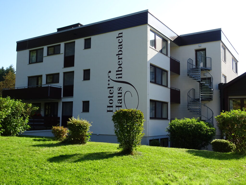 Außenansicht des mehrstöckigen weißen Hotels mit dem vertikal stehenden Schriftzug Hotel Silberbach, grüne Rasenflächen und eine spiralige Außentreppe an der Fassadenseite