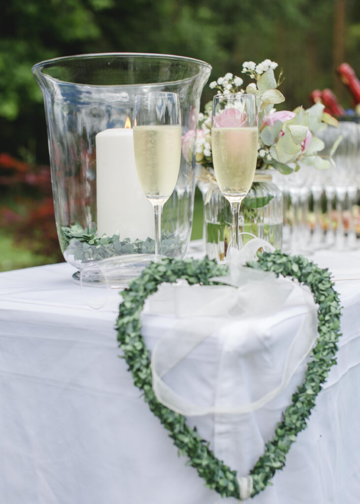 Sektempfang Dekoration mit Sektgläsern, einer großen Kerze in einer Glasvase und grünem Herzkranz auf einem weißen Tisch, vor grünem Garten – Hotel Silberbach Hochzeiten