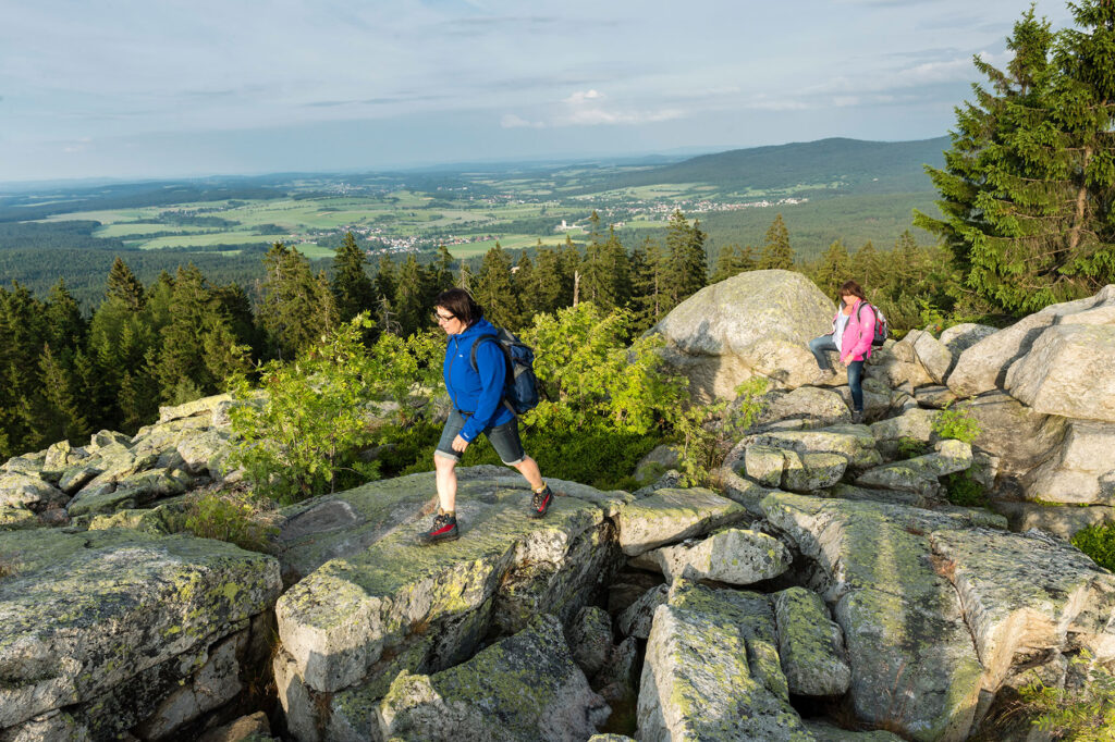 Wanderer im Fichtelgebirge: Eine Person in blauer Jacke klettert über Granitfelsen, im Hintergrund eine weitere Person mit rosa Jacke, Waldlandschaft und Blick ins Egertal
