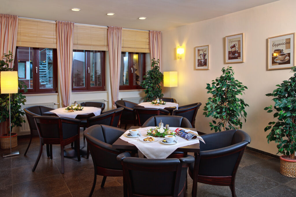 Coffeeshop-Innenraum des Hauses Silberbach in Selb mit hellen Wänden, dunklen Holztischen, Pflanzen und Fensterfronten; Tischdekorationen, Frühstückssets und Café-Atmosphäre.