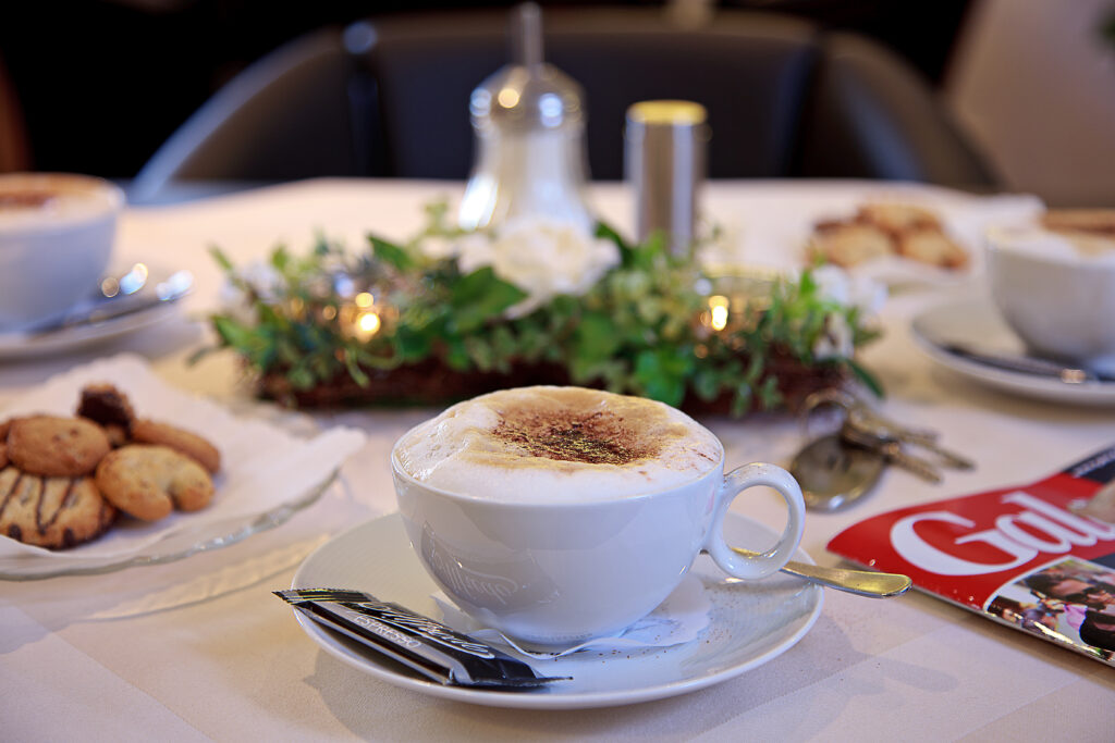 Eine weiße Kaffeetasse mit Milchschaum (Cappuccino) im Vordergrund, daneben Kekse und ein Blüten- und Kräutergesteck auf einem gedeckten Tisch – Verpflegung im Haus Silberbach, Selb.