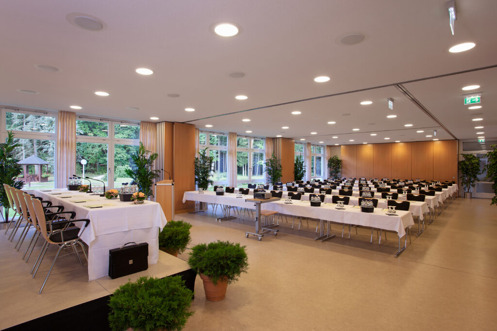 Konferenzraum im Haus Silberbach mit großen Fensterfronten, langen Tischen, weißen Tischdecken, Stühlen in Reihen, Grünpflanzen und moderner Beleuchtung