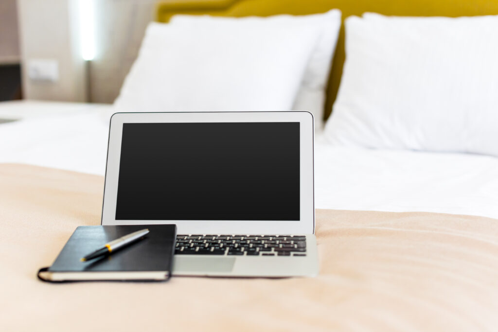 Silbernes Laptop auf beigefarbenem Bettlaken im Hotelzimmer, daneben schwarz gebundene Notizbuch mit Stift; ruhiges Ambiente im Hotel Haus Silberbach