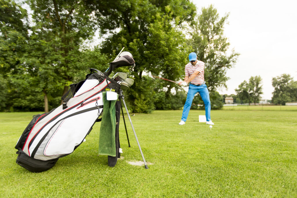 Golfer beim Abschlag auf einer grünen Golfwiese; Vordergrund mit einem weißen Golfbag, dahinter ein Mann in blau-gestreifter Hose und gestreiftem Oberteil, umgeben von Bäumen; Bezug zu Haus Silberbach und Naturpark Fichtelgebirge