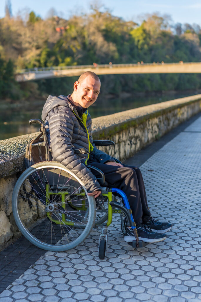Mann im Rollstuhl sitzt an einer Flussufer-Promenade mit Steinmauer, nahe dem Haus Silberbach; im Hintergrund Brücke und Herbstbäume, sonniger Tag.